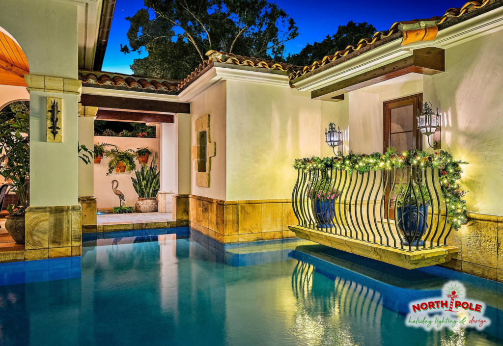 Custom pre-lit holiday garland draped over a luxury pool balcony overlooking a waterfront estate in Palm Beach.