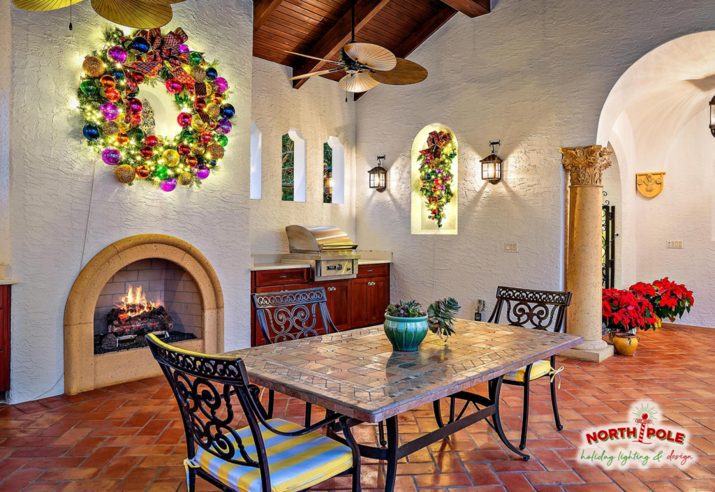 Luxury Boynton Beach patio featuring custom holiday decor with a large multi-color lit wreath and matching teardrop spritz over an outdoor fireplace.