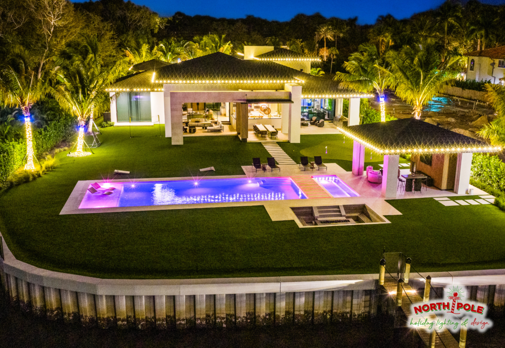 Aerial view of a luxury Jupiter backyard featuring warm white LED palm wraps with blue crowns, matching roofline lighting on the main house and pool house.